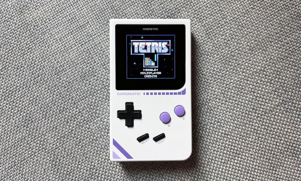The ModRetro Chromatic gaming handheld rests on a gray couch cushion, displaying the start screen to the video game Tetris.