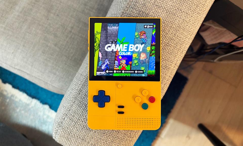 The Retroid Pocket Classic rests on the arm of a gray couch with its screen on displaying a Game Boy Color logo.